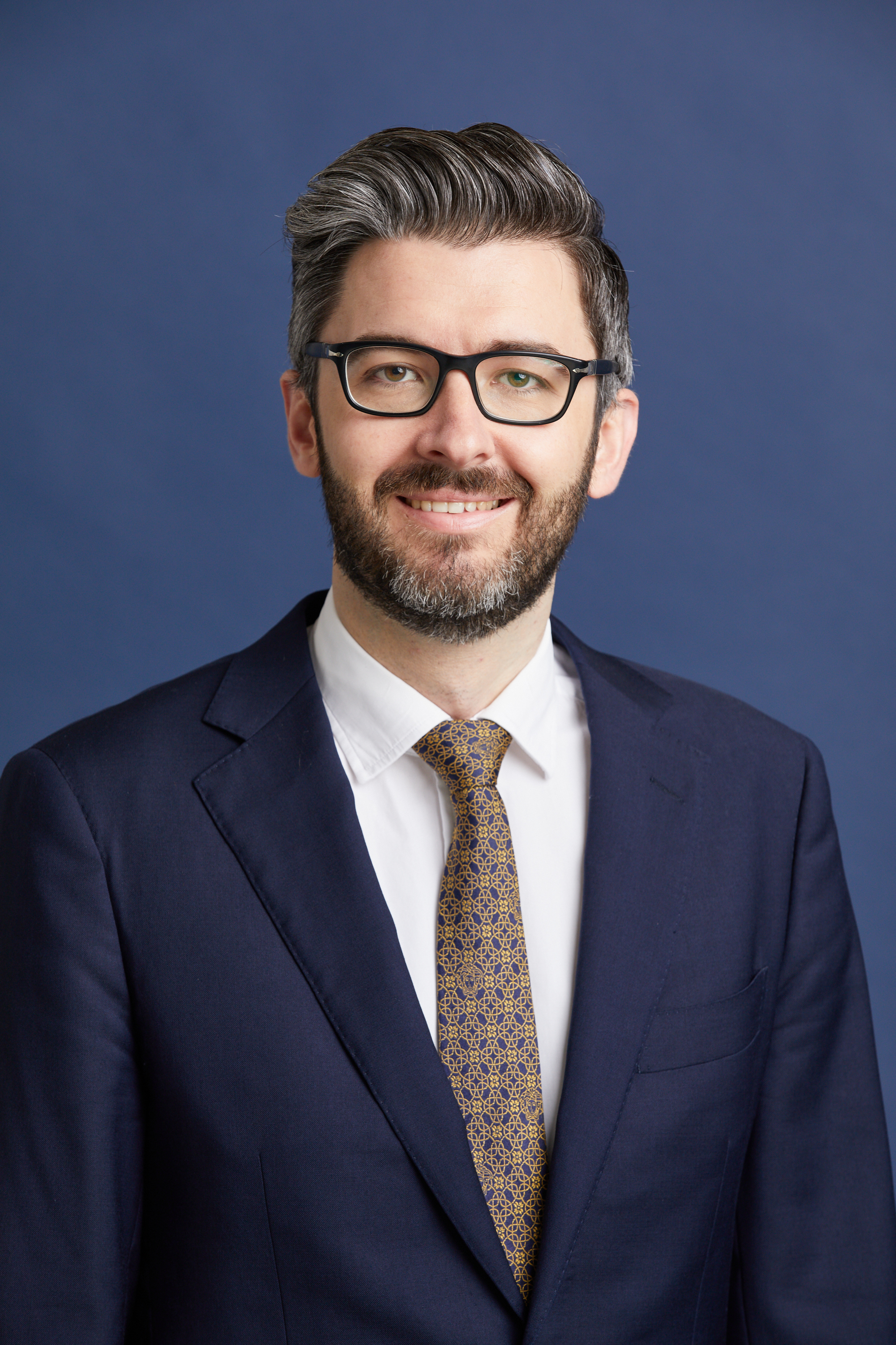Curtis Ward - a person (he/him) with glasses, a beard and smiling, wearing a navy blue suit and tie